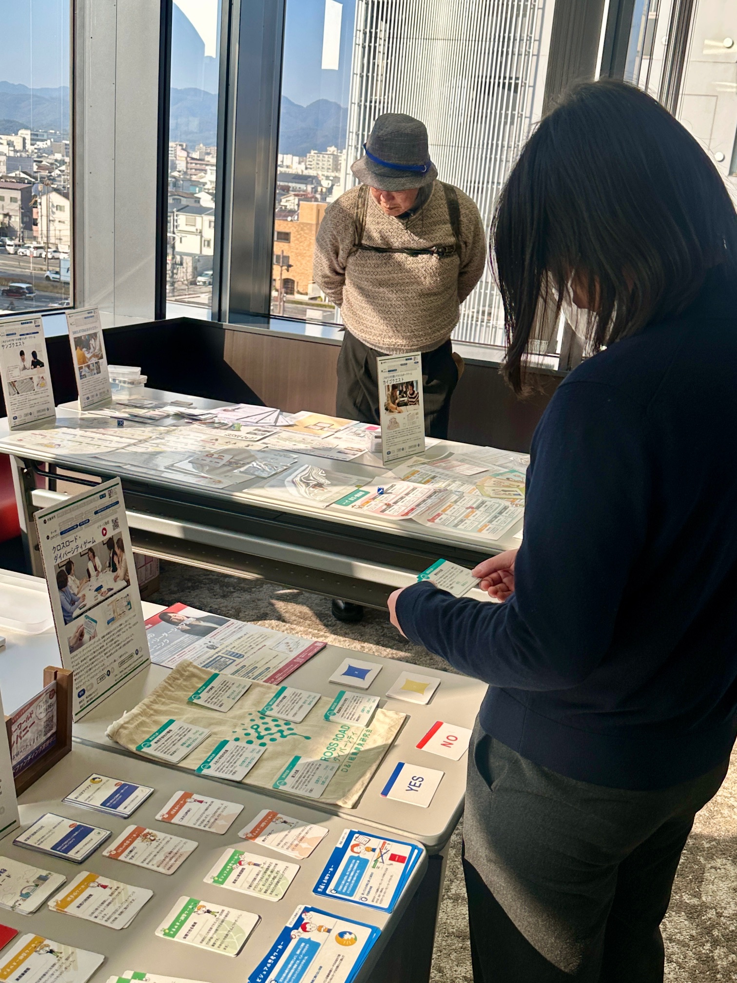 京都SDGsゲームショウの展示ブースにて、複数の来場者が「クロスロード・ダイバーシティゲーム」のカードやパンフレットを手に取り確認しています。DEIコンサルティングやダイバーシティ研修における実践的なツールとして、参加者の深い関心と理解を促しています。